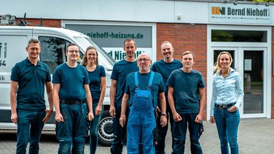 Die Mitarbeiter einer Heizungsfirma stehen in einer Reihe vor ihrem Büro, der ältere Mann in blauer Latzhose sticht hervor.