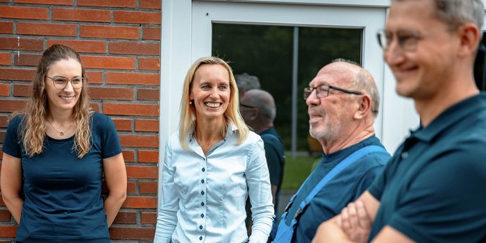 Eine Gruppe von vier Personen steht vor einer Backsteinwand, alle lächeln und scheinen sich gut zu unterhalten.