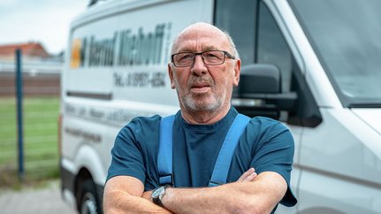 Mit verschränkten Armen und einem nachdenklichen Blick steht der Mann vor einem Lieferwagen, der seine Firma bewirbt.