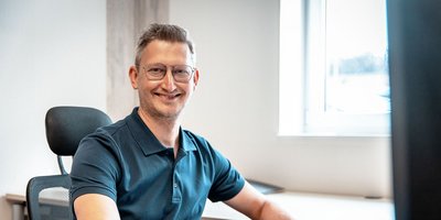 Der Mann mit kurzen Haaren und Brille strahlt Zufriedenheit aus, während er in einem modernen Arbeitsumfeld sitzt.